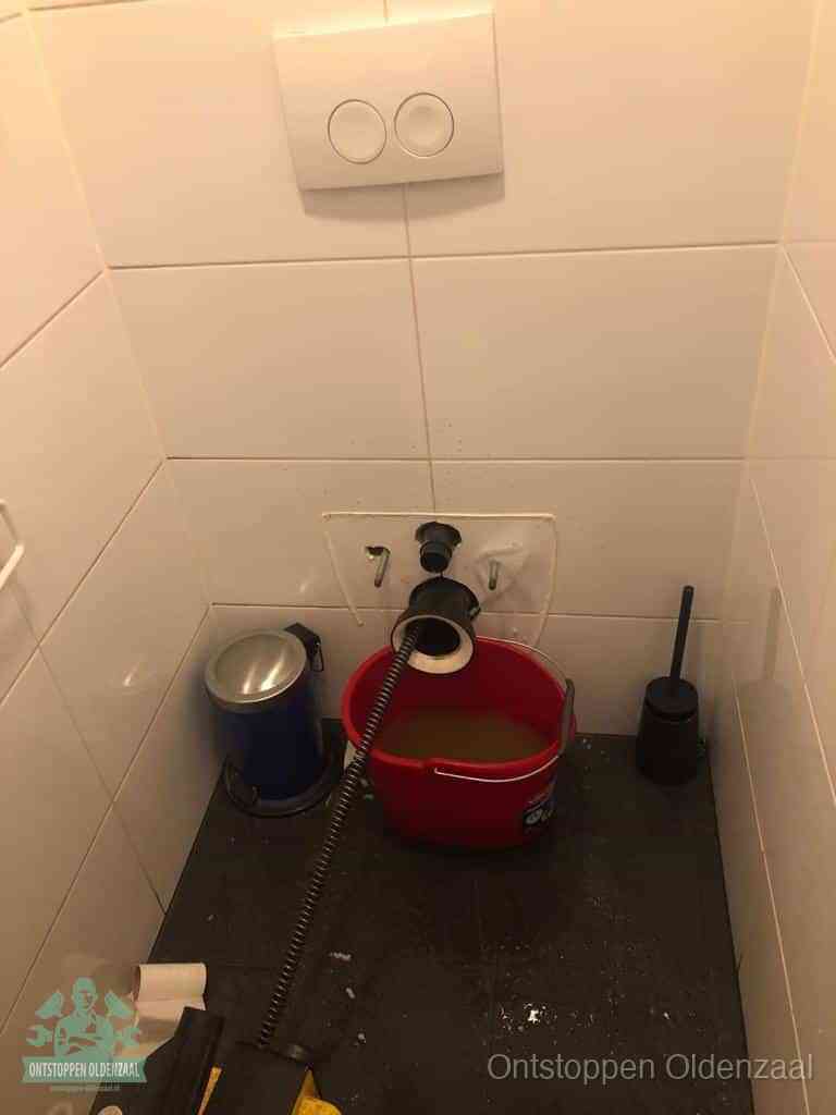 Een ontstopte wand-wc met verwijderde toiletpot waar een ontstoppingsveer in de wanduitgang verdwijnt, het achterhalen waar de verstopping zit in Oldenzaal