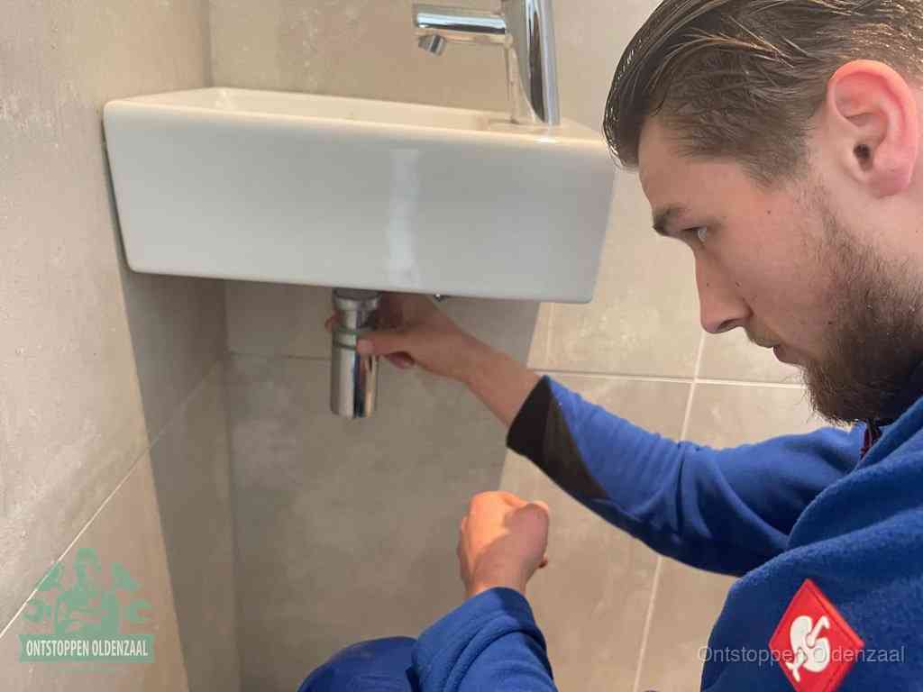 Een monteur draait met de hand de zwanenhalssifon onder een fonteinwasbak in de toiletruimte los om de bron van rioollucht in Oldenzaal te vinden