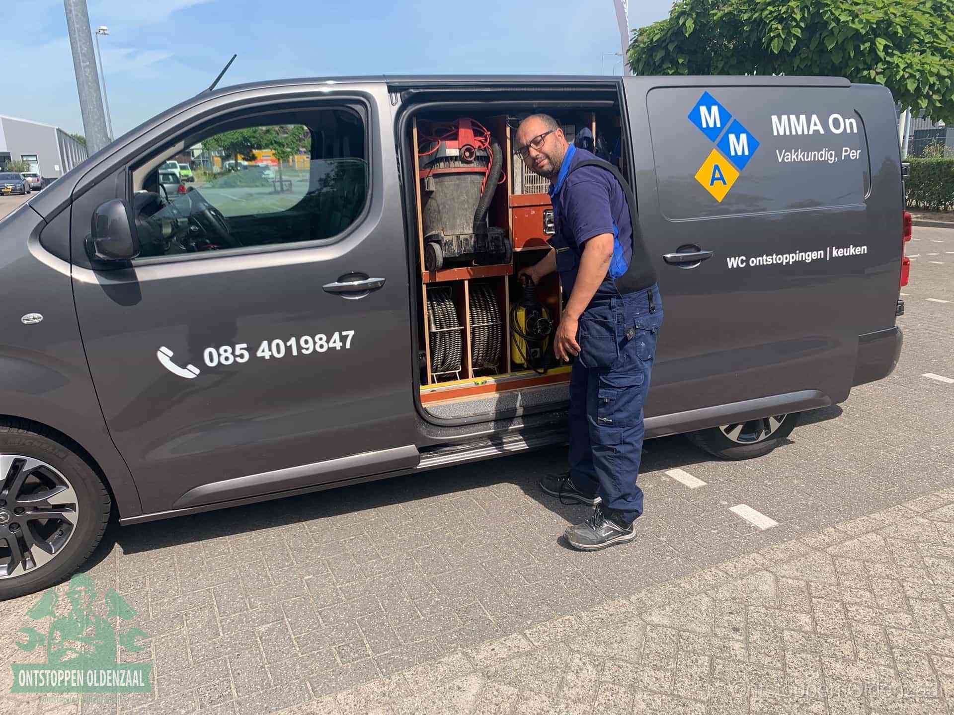 Een monteur bij de open schuifdeur van zijn bedrijfsbus op een parkeerplaats, klaar om de oorzaak van terugkerende afvoerverstoppingen aan te pakken in Oldenzaal