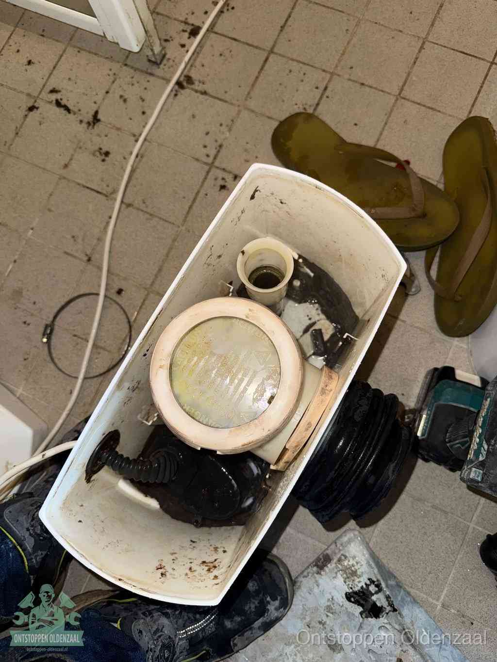 Een gedemonteerde toiletpot op de vloer met ontstoppingsveer en gereedschap ernaast, het verwijderen van een vastzittend voorwerp uit de wc in Oldenzaal